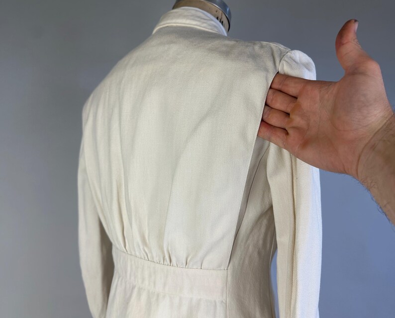 May include: An off-white jacket featuring a structured collar, long sleeves, and a fitted waistline. The back panel displays vertical pleats that extend from the shoulder down to the waist seam, creating a tailored silhouette with a clean finish.