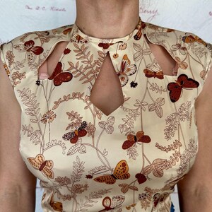 May include: Cream-colored sleeveless top with a butterfly and floral pattern. The top features cut-out details at the shoulders and neckline. The butterflies are in shades of brown and orange, with brown floral accents.