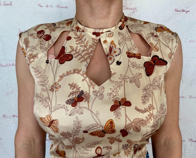 May include: Cream-colored sleeveless top with a butterfly and floral pattern. The top features cut-out details at the shoulders and neckline. The butterflies are in shades of brown and orange, with brown floral accents.