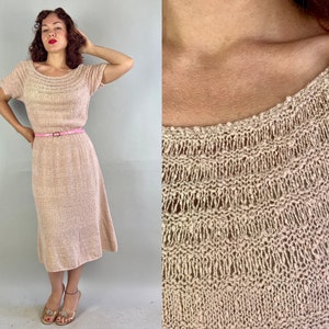May include: A light pink knitted dress with short sleeves and a midi-length skirt. The dress has a round neckline and is cinched at the waist with a pink belt. The fabric has a textured appearance. The dress is a vintage style.