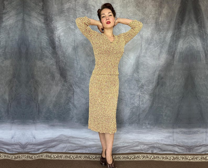 May include: A vintage two-piece knit dress in a textured, speckled pattern. The set includes a long-sleeved top with a V-neck and a knee-length pencil skirt. The dress is a mix of yellow and brown tones, and the model is wearing brown heels.