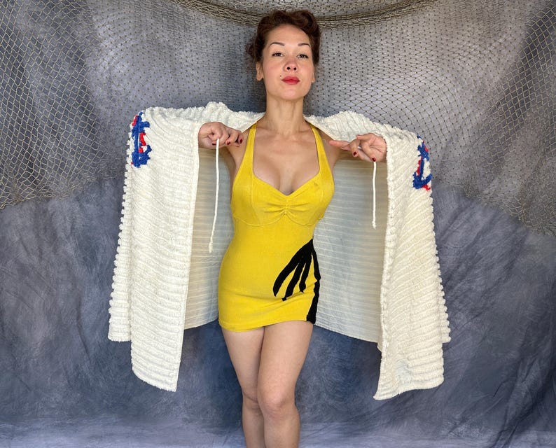 May include: A yellow sleeveless mini dress with a sweetheart neckline and a black graphic design on the side, paired with a cream-colored textured knit cardigan featuring blue and red crochet accents on the shoulders and white drawstring ties.