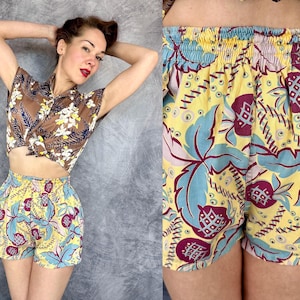 Op de afbeelding: Een tweedelige outfit met een bruine bloemen croptop en een high-waisted short. De short heeft een gele achtergrond met een herhalend patroon van blauwe bladeren en kastanjebruine vruchten. De top is bruin met witte en gele bloemmotieven.