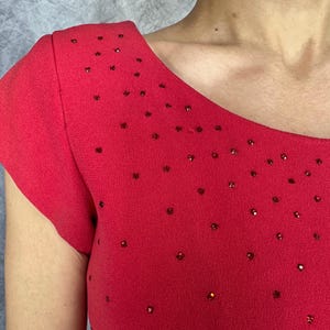 May include: A bright red short-sleeved top featuring a wide, rounded neckline. The fabric is adorned with numerous small, scattered red rhinestones that create a subtle, sparkling effect across the chest and shoulder areas of the garment.
