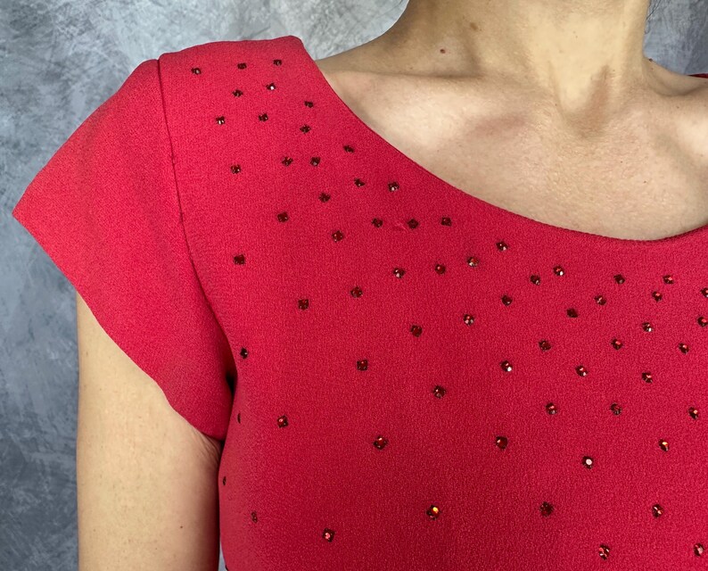 May include: A bright red short-sleeved top featuring a wide, rounded neckline. The fabric is adorned with numerous small, scattered red rhinestones that create a subtle, sparkling effect across the chest and shoulder areas of the garment.