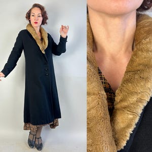 Op de afbeelding: Een vintage zwarte jas met een imitatiebontkraag. De lange jas heeft een knoopsluiting aan de voorkant en lange mouwen. De kraag is lichtbruin van kleur. De jas is een klassieke stijl, geschikt voor kouder weer.