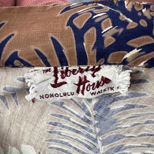 Może przedstawiać: Zbliżenie na tkaninę w stylu vintage z tropikalnym wzorem liści w odcieniach brązu, niebieskiego i beżu. Tkanina posiada metkę z napisem "The Liberty House Honolulu Waikiki."