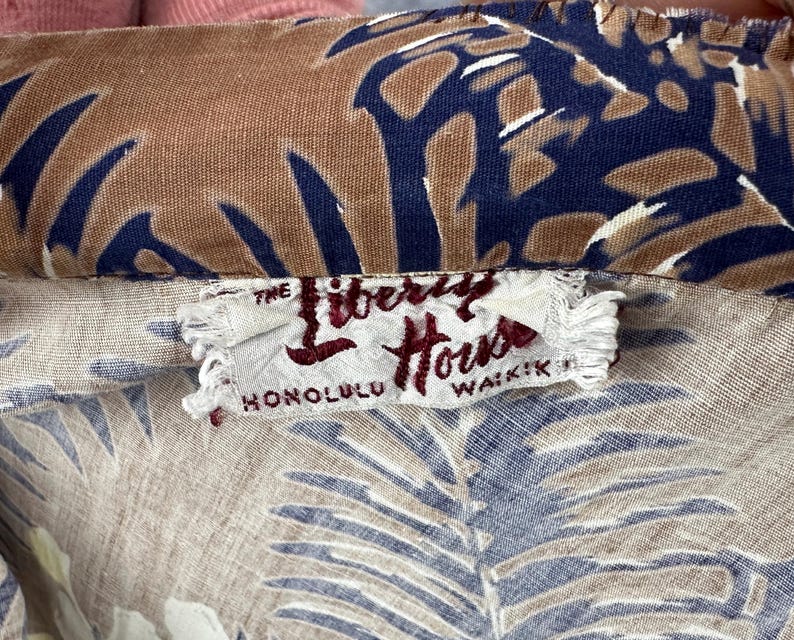 Może przedstawiać: Zbliżenie na tkaninę w stylu vintage z tropikalnym wzorem liści w odcieniach brązu, niebieskiego i beżu. Tkanina posiada metkę z napisem "The Liberty House Honolulu Waikiki."
