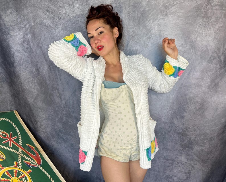 May include: A white textured cardigan featuring long sleeves with colorful floral crochet accents in yellow, blue, and pink. The cardigan has two front pockets with matching floral details and is worn over a light yellow polka dot romper.
