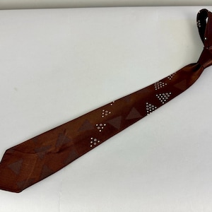 Może przedstawiać: Brązowy krawat z powtarzającym się wzorem trójkątów i białych kropek. Krawat jest zawiązany u góry i leży po przekątnej na białej powierzchni. Tkanina krawata ma subtelny połysk.