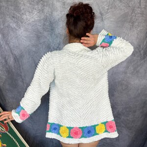 May include: A white long-sleeved jacket with a textured, chevron-patterned surface. The cuffs and bottom hem feature a teal band decorated with a repeating pattern of three-dimensional fabric roses in shades of pink, blue, and yellow.