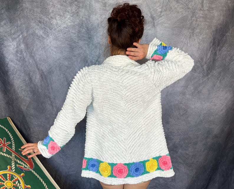 May include: A white long-sleeved jacket with a textured, chevron-patterned surface. The cuffs and bottom hem feature a teal band decorated with a repeating pattern of three-dimensional fabric roses in shades of pink, blue, and yellow.