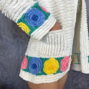 May include: A white textured terry cloth jacket featuring a long sleeve with a cuff decorated in blue, yellow, and pink floral appliques. The hem of the jacket also displays a horizontal band of blue, yellow, and pink circular floral designs.