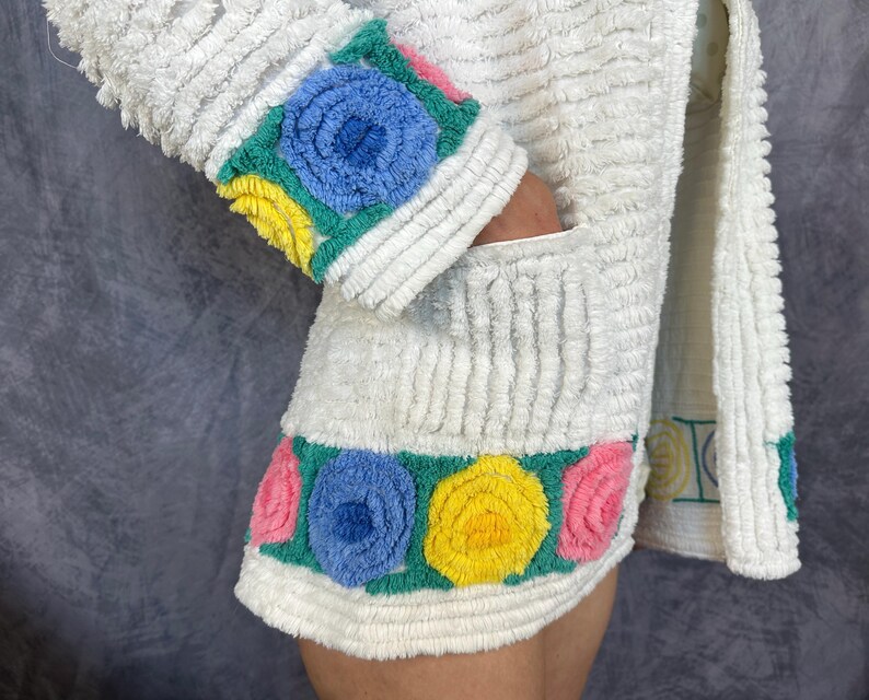 May include: A white textured terry cloth jacket featuring a long sleeve with a cuff decorated in blue, yellow, and pink floral appliques. The hem of the jacket also displays a horizontal band of blue, yellow, and pink circular floral designs.