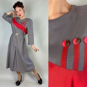 Könnte beinhalten: Ein Vintage-Kleid in Grau mit einem roten Akzent über der Brust und einem passenden Gürtel. Das Kleid hat drei rote vertikale Streifen mit goldfarbenen Knöpfen. Das Kleid hat lange Ärmel und einen knielangen Rock.
