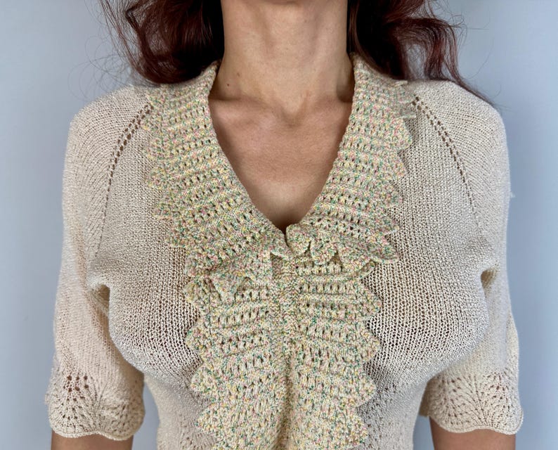 May include: A cream-colored crocheted sweater with a scalloped, multi-colored trim around the neckline.