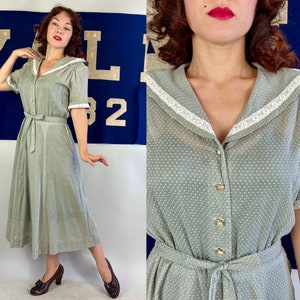 May include: A vintage-style sage green dress with white polka dots. The dress features a sailor collar with white lace trim, short sleeves, and a matching belt. Gold-colored buttons run down the front. The dress is knee-length and has a flowing skirt.