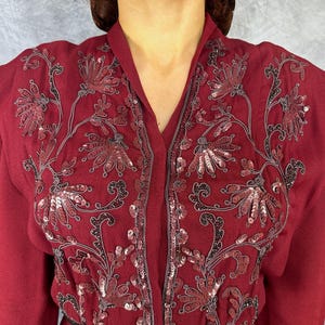 May include: A deep burgundy jacket with detailed floral embroidery and sequin accents. The jacket has a V-neck and long sleeves. The embroidery uses dark grey thread, providing a contrast against the burgundy material. The design is elegant and detailed.