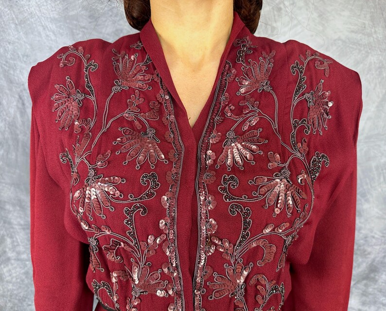 May include: A deep burgundy jacket with detailed floral embroidery and sequin accents. The jacket has a V-neck and long sleeves. The embroidery uses dark grey thread, providing a contrast against the burgundy material. The design is elegant and detailed.