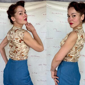 May include: A sleeveless, cream-colored top with a butterfly pattern and a mandarin collar. The top is paired with high-waisted, blue denim pants. The outfit is vintage-inspired, with a retro aesthetic. The background is a white fabric with handwritten text.