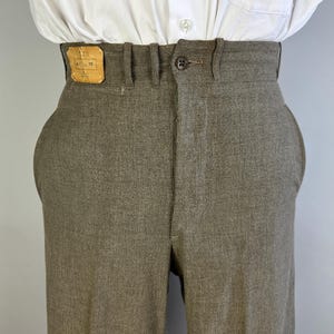 Op de afbeelding: Een taupekleurige broek met hoge taille, voorzien van een enkele donkerbruine knoopsluiting, riemlussen en een rechthoekige lichtbruine leren patch op de tailleband aan de voorzijde. De broek wordt gedragen met een wit overhemd.