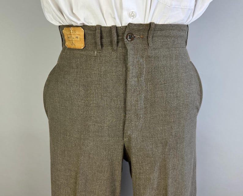 Op de afbeelding: Een taupekleurige broek met hoge taille, voorzien van een enkele donkerbruine knoopsluiting, riemlussen en een rechthoekige lichtbruine leren patch op de tailleband aan de voorzijde. De broek wordt gedragen met een wit overhemd.
