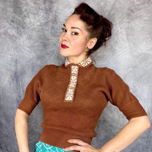 Pu&ograve; includere: Un maglione marrone a maniche corte con colletto e pannello frontale decorativi. Il colletto e il pannello presentano un design geometrico bianco e beige. Il maglione ha una trama a coste e una silhouette aderente. Lo sfondo &egrave; grigio screziato.