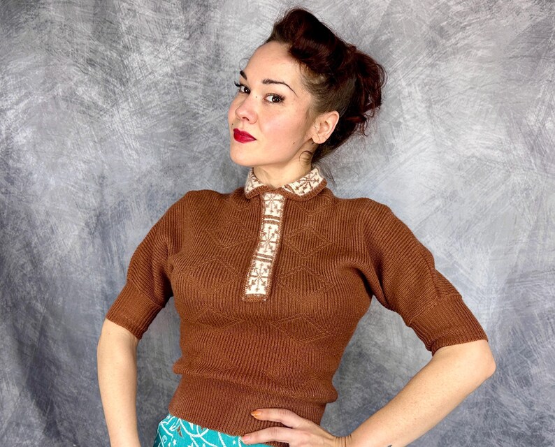 Pu&ograve; includere: Un maglione marrone a maniche corte con colletto e pannello frontale decorativi. Il colletto e il pannello presentano un design geometrico bianco e beige. Il maglione ha una trama a coste e una silhouette aderente. Lo sfondo &egrave; grigio screziato.