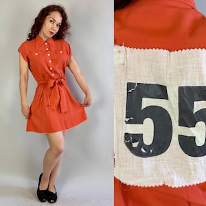 May include: A vintage orange dress with a collared neckline, short sleeves, and a matching tie belt. The dress has white buttons and a white hem. The back of the dress features a white patch with the number "55" in black.