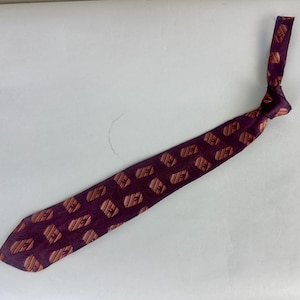 May include: A purple necktie with a repeating pattern of orange and brown geometric shapes. The tie is knotted and lies on a white surface. The tie is a classic accessory.