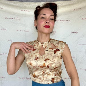 May include: A cream-colored sleeveless top with a keyhole neckline and a butterfly and floral pattern in shades of brown and gold. The top is made of a silky material.