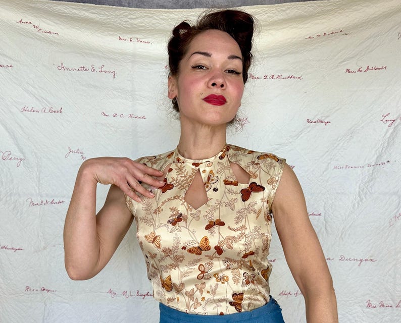 May include: A cream-colored sleeveless top with a keyhole neckline and a butterfly and floral pattern in shades of brown and gold. The top is made of a silky material.