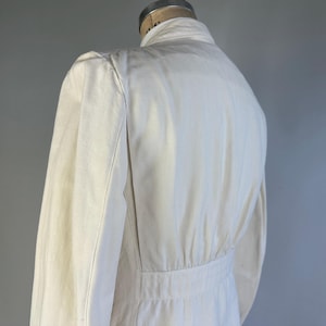 May include: An off-white jacket featuring a structured collar, long sleeves, and a fitted waistline with vertical paneling. The garment is displayed on a mannequin, showcasing a clean, solid-coloured design with visible seams along the shoulders and back.