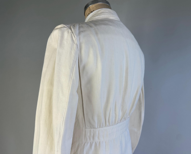 May include: An off-white jacket featuring a structured collar, long sleeves, and a fitted waistline with vertical paneling. The garment is displayed on a mannequin, showcasing a clean, solid-coloured design with visible seams along the shoulders and back.