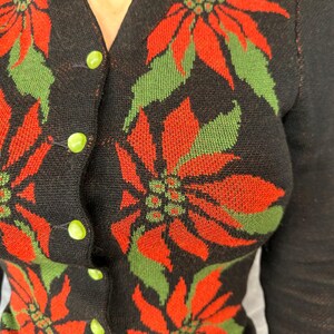 May include: Black cardigan sweater with a poinsettia pattern in red and green. The sweater has a V-neck and is fastened with lime green buttons. The sleeves are long and the overall style is vintage.