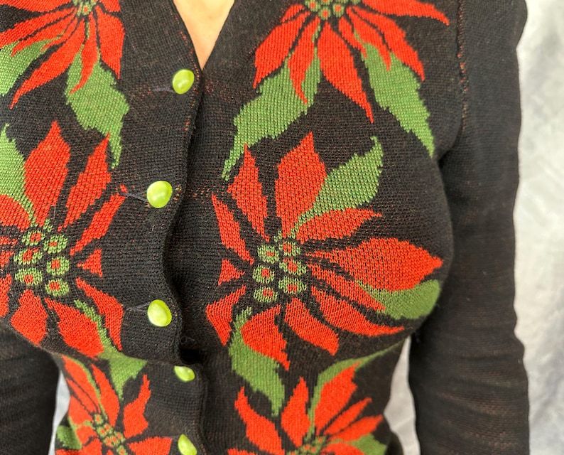 May include: Black cardigan sweater with a poinsettia pattern in red and green. The sweater has a V-neck and is fastened with lime green buttons. The sleeves are long and the overall style is vintage.