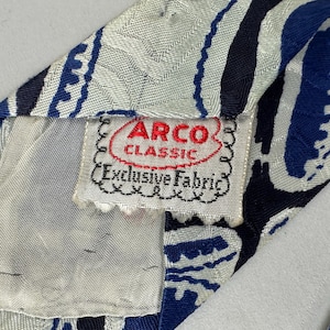 May include: A necktie featuring a white, navy blue, and royal blue abstract pattern with a textured fabric finish. A white fabric label is stitched to the back, displaying the red and black text ARCO CLASSIC and Exclusive Fabric.