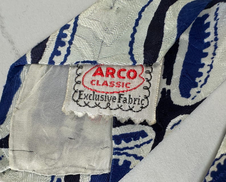 May include: A necktie featuring a white, navy blue, and royal blue abstract pattern with a textured fabric finish. A white fabric label is stitched to the back, displaying the red and black text ARCO CLASSIC and Exclusive Fabric.