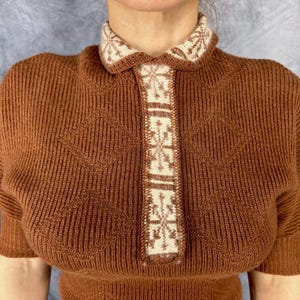 Pu&ograve; includere: Top in maglia marrone con colletto e pannello centrale color crema. Il colletto e il pannello presentano un motivo geometrico ripetuto. Il top a maniche corte ha una trama a coste e una silhouette aderente, adatta per il clima fresco.