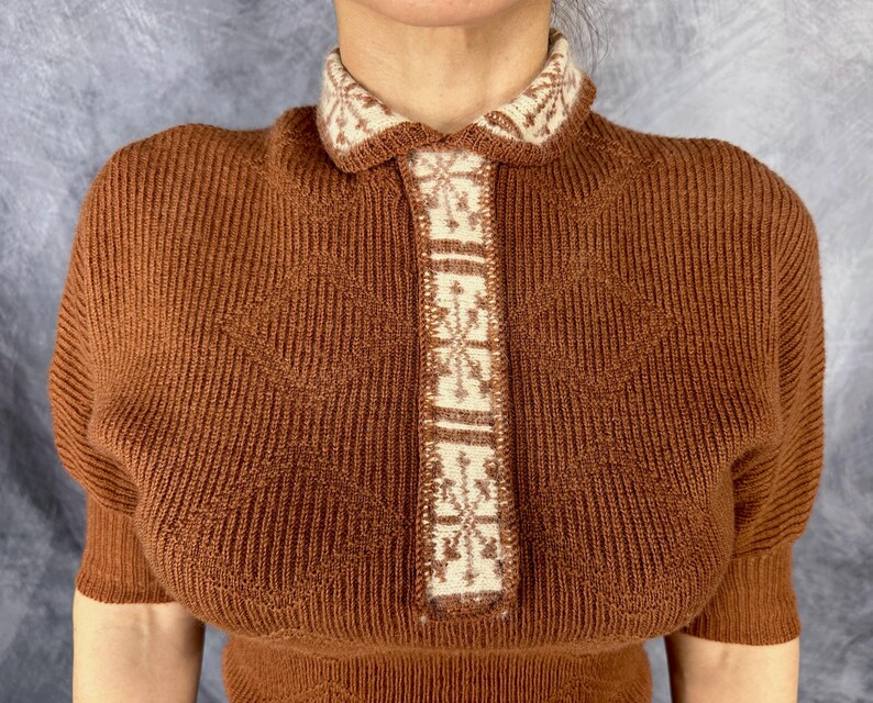 Pu&ograve; includere: Top in maglia marrone con colletto e pannello centrale color crema. Il colletto e il pannello presentano un motivo geometrico ripetuto. Il top a maniche corte ha una trama a coste e una silhouette aderente, adatta per il clima fresco.