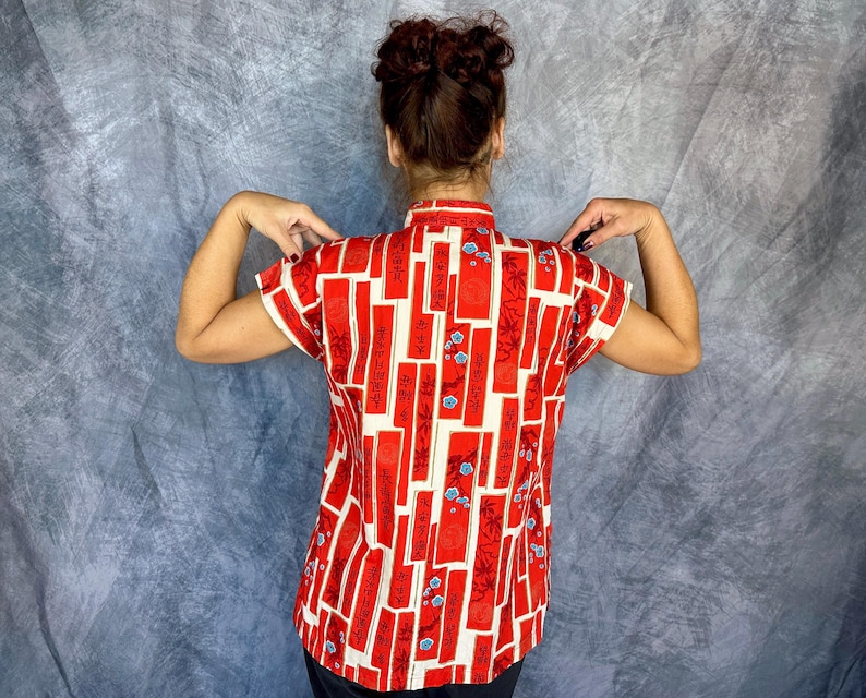 Pode incluir: Uma camisa de manga curta com gola mandarim e um padr&atilde;o vibrante de blocos retangulares vermelhos intercalados com sec&ccedil;&otilde;es brancas. O design inclui caracteres de caligrafia preta e pequenos detalhes florais azuis por toda a pe&ccedil;a.