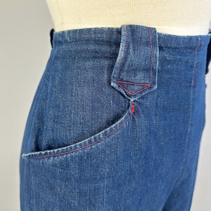 May include: High-waisted dark blue denim pants featuring a prominent belt loop with a pointed bottom edge. The garment is detailed with contrasting red stitching along the pocket opening, the belt loop, and the side seams, showcasing a structured fit.