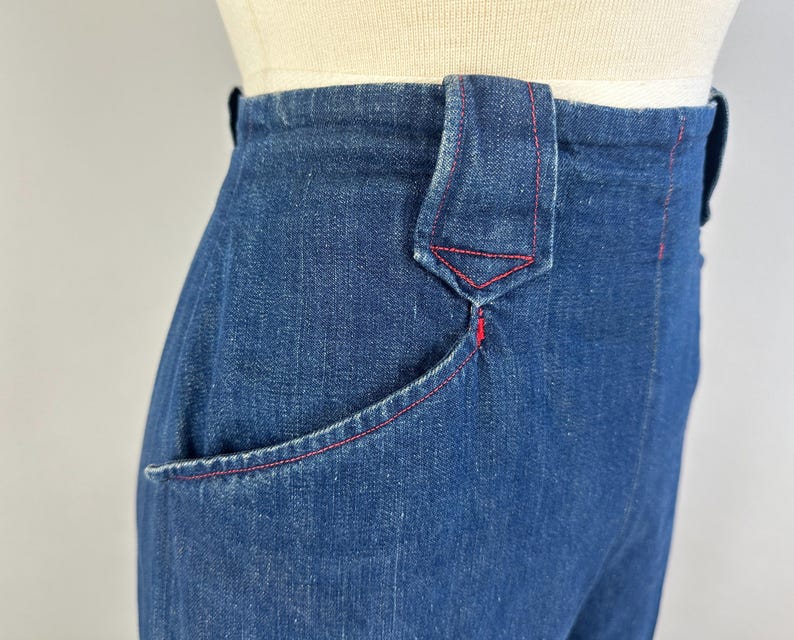 May include: High-waisted dark blue denim pants featuring a prominent belt loop with a pointed bottom edge. The garment is detailed with contrasting red stitching along the pocket opening, the belt loop, and the side seams, showcasing a structured fit.