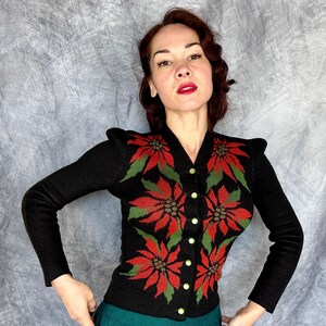 May include: Black cardigan sweater with a V-neck and long sleeves. The sweater features a poinsettia floral design in red and green. The sweater has gold-colored buttons.