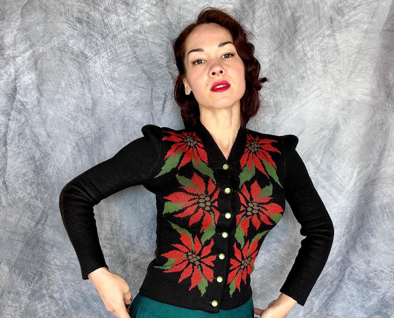 May include: Black cardigan sweater with a V-neck and long sleeves. The sweater features a poinsettia floral design in red and green. The sweater has gold-colored buttons.