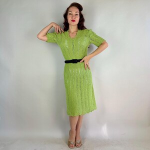 May include: A lime green, short-sleeved dress with a V-neck and a black belt. The dress has a textured, open-knit design and a scalloped hemline. The dress is knee-length.