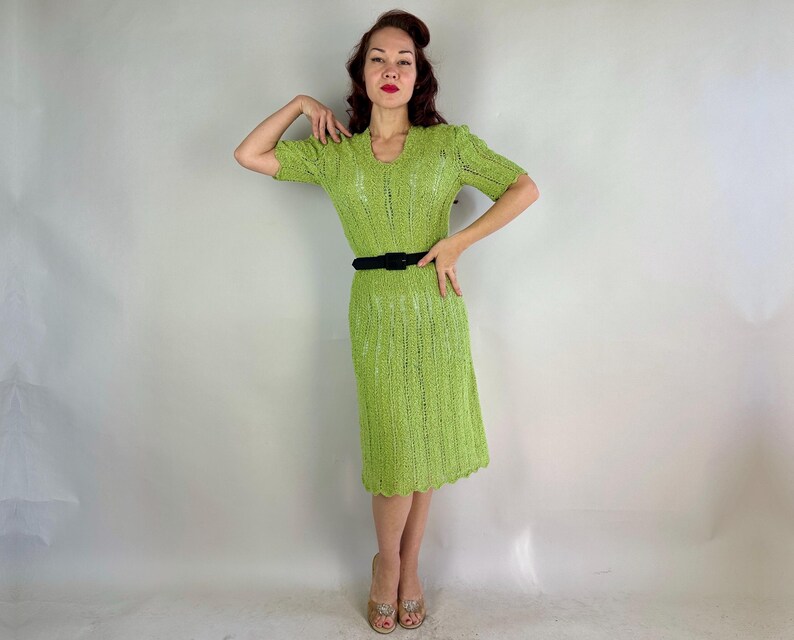 May include: A lime green, short-sleeved dress with a V-neck and a black belt. The dress has a textured, open-knit design and a scalloped hemline. The dress is knee-length.