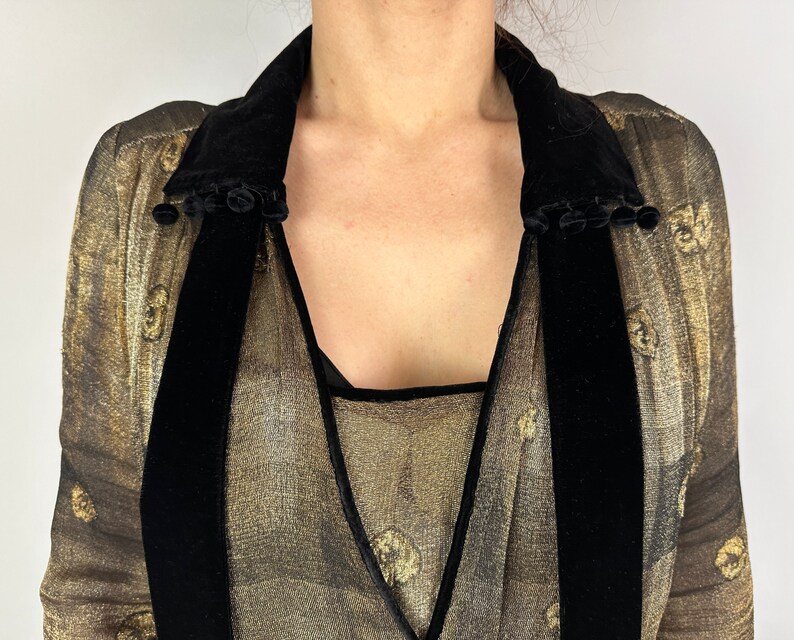 May include: A gold sheer blouse with a black velvet collar and trim. The collar has a row of black pom-poms along the edge.