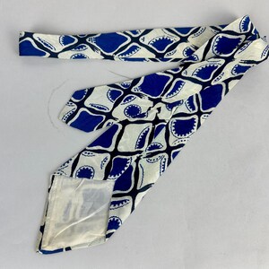 May include: A necktie featuring a repeating geometric pattern of blue and white shapes. The design consists of interconnected blue diamond-like forms with scalloped edges against a cream-colored background. The fabric has a smooth, satin-like finish.