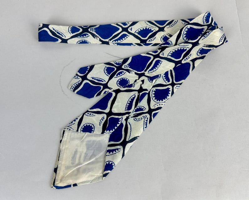 May include: A necktie featuring a repeating geometric pattern of blue and white shapes. The design consists of interconnected blue diamond-like forms with scalloped edges against a cream-colored background. The fabric has a smooth, satin-like finish.